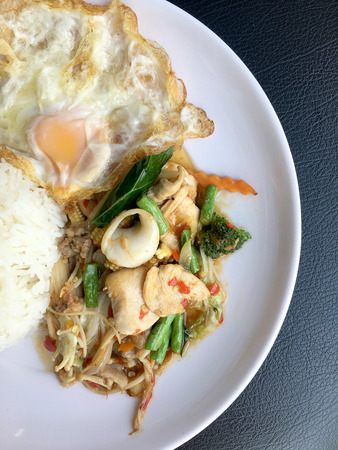 Fried spicy fried seafood with basil leaves and mixed vegetable and fried egg on jasmine rice in white dish. Thai style food.の写真素材