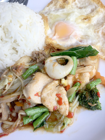 Fried spicy fried seafood with basil leaves and mixed vegetable and fried egg on jasmine rice in white dish. Thai style food.の写真素材