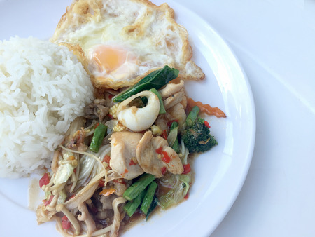 Fried spicy fried seafood with basil leaves and mixed vegetable and fried egg on jasmine rice in white dish. Thai style food.の写真素材
