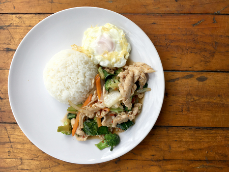 stir-fried chicken with mixed vegetables and fried egg in thai style with rice in white dish on old wooden table. Asian thai food.の写真素材