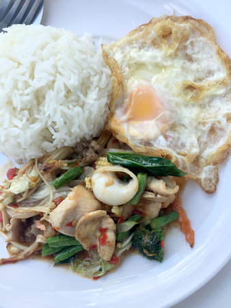 Fried spicy fried seafood with basil leaves and mixed vegetable and fried egg on jasmine rice in white dish. Thai style food.の写真素材