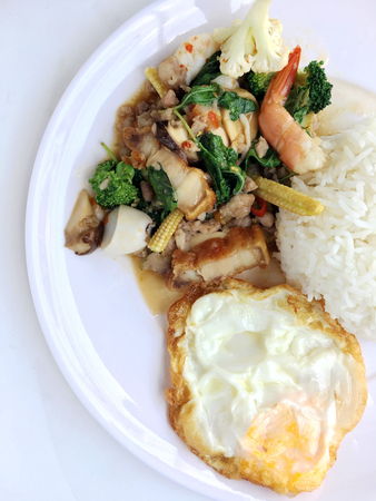 stir fried crispy pork, shrimp, mix seafood with thai holy basil, chilies, garlic, and soy sauce. in white dish with rice and thai fried egg style topping on white background.の写真素材