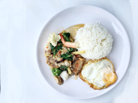 stir fried crispy pork, shrimp, mix seafood with thai holy basil, chilies, garlic, and soy sauce. in white dish with rice and thai fried egg style topping on white background.の写真素材