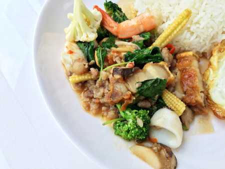 stir fried crispy pork, shrimp, mix seafood with thai holy basil, chilies, garlic, and soy sauce. in white dish with rice and thai fried egg style topping on white background.の写真素材