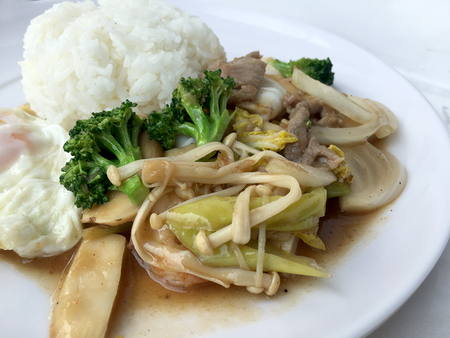 Thai cuisine. Stir-fried colorful vegetables, onion, broccoli, mushroom with meat in white plate with rice and fried egg on white background. Thai style food in fast street restaurant.の写真素材