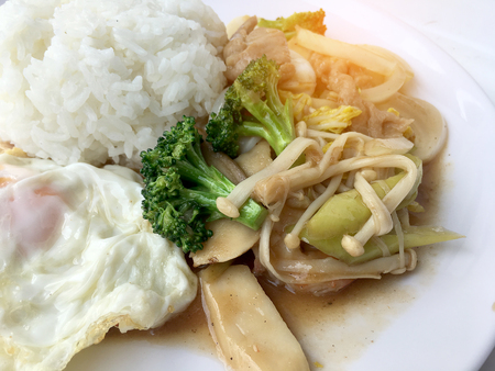 Thai cuisine. Stir-fried colorful vegetables, onion, broccoli, mushroom with meat in white plate with rice and fried egg on white background. Thai style food in fast street restaurant.の写真素材