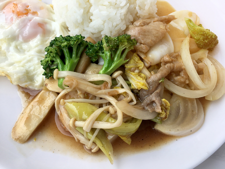 Thai cuisine. Stir-fried colorful vegetables, onion, broccoli, mushroom with meat in white plate with rice and fried egg on white background. Thai style food in fast street restaurant.の写真素材