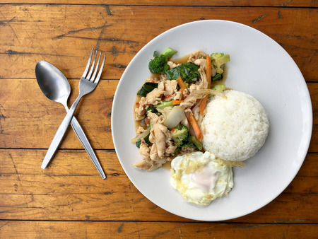 stir-fried chicken with mixed vegetables and fried egg in thai style with rice in white dish on old wooden table. Asian thai food.の写真素材