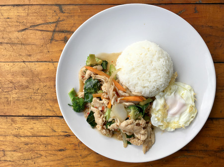 stir-fried chicken with mixed vegetables and fried egg in thai style with rice in white dish on old wooden table. Asian thai food.の写真素材