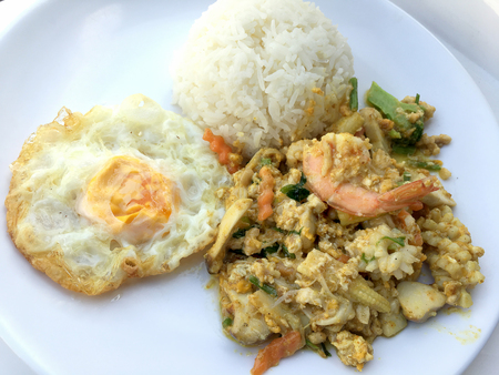 Stir-fried seafood, prawn and squid with yellow curry powder with rice and fried egg in white plate on white background. Thai style food.の写真素材