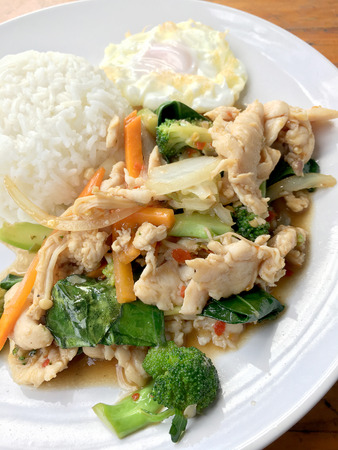 stir-fried chicken with mixed vegetables and fried egg in thai style with rice in white dish on old wooden table. Asian thai food.の写真素材