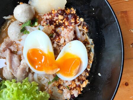 Thai spicy pork noodle soup with coconut milk, vegetables herbs and onsen tamago eggs  in black bowl with sesame seeds and peanut on wooden table, view from aboveの写真素材
