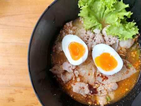 Thai spicy pork noodle soup with coconut milk, vegetables herbs and onsen tamago eggs  in black bowl with sesame seeds and peanut on wooden table, view from aboveの写真素材
