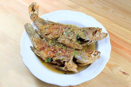 Thai Giant gourami fish and garlics deep fried and sweet spicy sauce in white dish on pine wooden table. Deep fried fish with Sweet chili. Thai food fried fish with spicy sauce. Thai style food.の写真素材