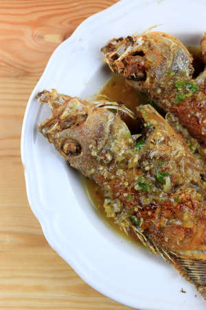 Thai Giant gourami fish and garlics deep fried and sweet spicy sauce in white dish on pine wooden table. Deep fried fish with Sweet chili. Thai food fried fish with spicy sauce. Thai style food.の写真素材