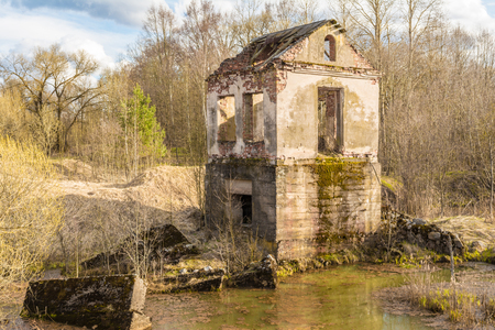 The old one is erected building of the hydroelectric plant with moss-grown-up walls is located in a forest surrounded by dry grass and shrubs, a sunny spring day, a blue sky with cloudsの写真素材
