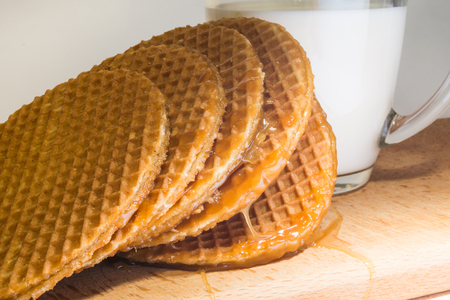 Round cookies or wafers with a layer of caramel lies on a rough pile on a wooden board, mug of clear glass with milk, food close-up abstract backgroundの写真素材