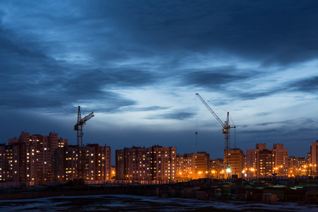 building site with two tower cranes against the background of multi-storey houses and a dark blue sky, night sceneの写真素材
