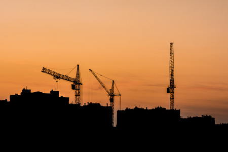 silhouettes of three tower cranes and houses during sunset, sunset with an orange sky in the cityの写真素材