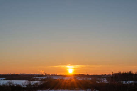 Winter evening landscape with sunset and multicolor sky. On the horizon is an urban industrial zone with smoking chimneys and a TV broadcaster. A view of the snow-covered area with dry grass, trees and bushes.の写真素材