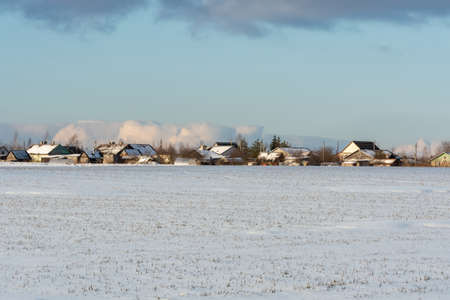 Snowy field with rare dry tava. On the horizon are the houses of a small village. Blue sky with a strip of clouds. Nature landscape backgroundの写真素材
