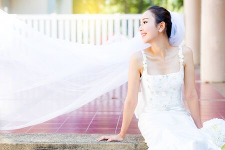 bride is sitting with flowers. Beautiful Young woman posing in park or garden in white bridal dress outdoors on a bright sunny day.の写真素材