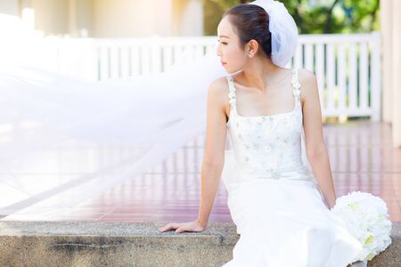 bride is sitting with flowers. Beautiful Young woman posing in park or garden in white bridal dress outdoors on a bright sunny day.の写真素材