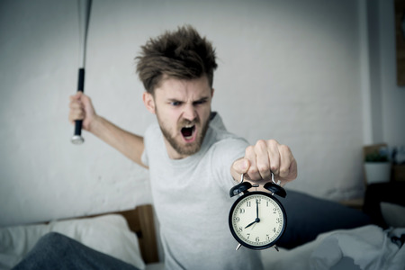 Portrait upset angry with baseball hit young man screaming at alarm clock on bedroom. Employee running late. Time management with business conceptの写真素材