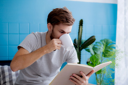 Young man reading book and drinking coffee on sofa in the living room, education conceptの写真素材