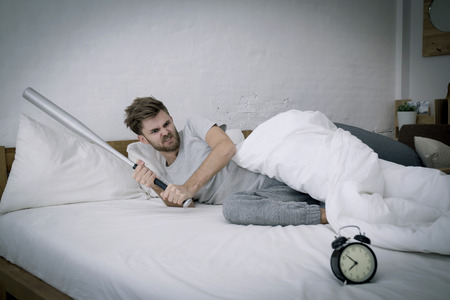 Portrait upset angry with baseball hit young man screaming at alarm clock on bedroom. Employee running late. Time management with business conceptの写真素材