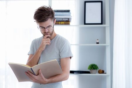 Handsome guy in eyeglasses is reading book preparing exam with standing at the living room, education conceptの写真素材