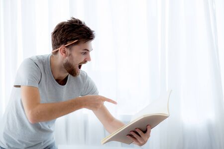 Handsome guy in eyeglasses is reading book preparing exam with standing at the living room, education conceptの写真素材