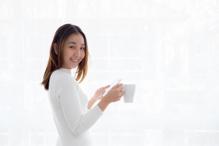 Beautiful young woman in white sweater drinking coffee and holding smartphone at a window.の写真素材
