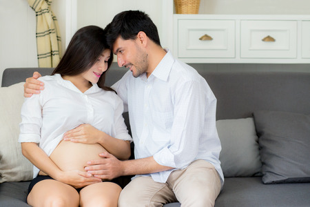 Beautiful pregnant asian woman and husband together care her pregnant on a sofa, Expectant parents,...