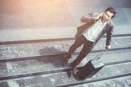 Asian elegant businessman wear suit talking mobile phone while walking holding suitcase outdoor on street, young man contact with customer appointment, communication business concept.の写真素材