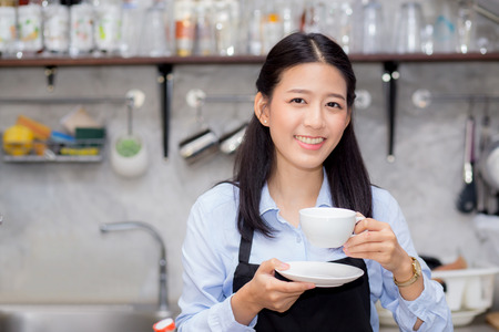 Portrait of beautiful young barista drinking a cup of coffee, asian woman is a employee standing in counter coffee shop, service concept.の写真素材