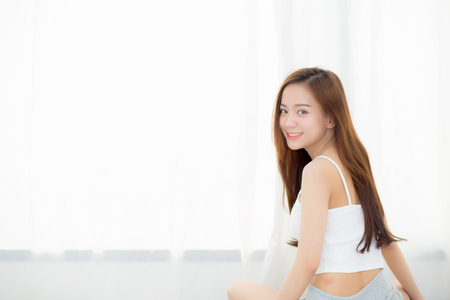 Beautiful asian young woman smiling with happiness leisure sitting on bed at home by the window, girl relax and wellness concept.の写真素材