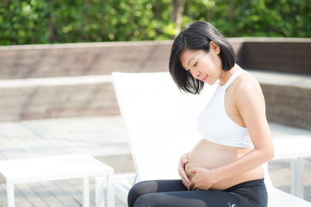 Portrait of beautiful pregnant asian young woman relax in the park, girl with belly sitting at swimming pool for wellness, tummy girl, health care concept.の写真素材