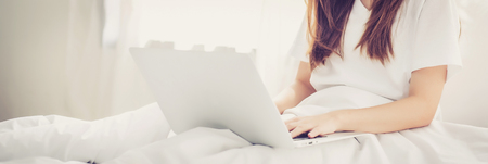 Banner website beautiful asian young woman setting on bed using laptop computer at bedroom for leisure and relax, freelance with girl working notebook, communication and lifestyle concept.の写真素材