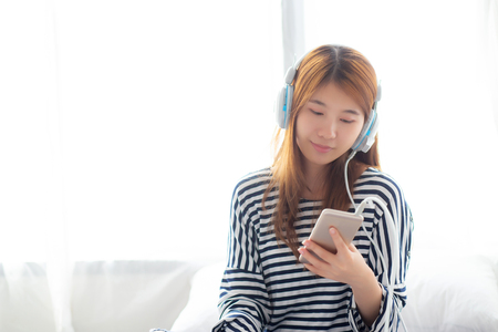 Beautiful asian young woman enjoy listen music with headphone and holding smart mobile phone while sitting in bedroom, relax girl with earphone, leisure and technology concept.の写真素材