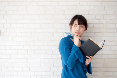 Beautiful asian woman smiling standing thinking and writing notebook on concrete cement white background at home, girl homework on book, education and lifestyle concept.の写真素材