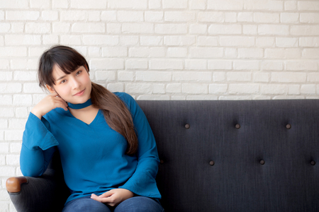 Beautiful portrait young asian woman sitting and smiling happy and looking at camera on sofa with casual at living room, girl cheerful and relax on couch at home, lifestyle concept.の写真素材
