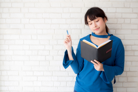 Beautiful asian woman smiling standing thinking and writing notebook on concrete cement white background at home, girl homework on book, education and lifestyle concept.の写真素材