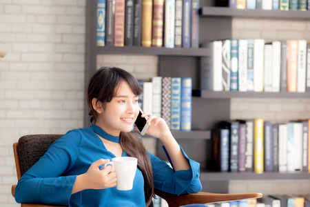 Beautiful portrait young asian woman smiling using mobile smart phone talking enjoy and drink coffee with relax, girl sitting on chair at living room, communication concept.の写真素材