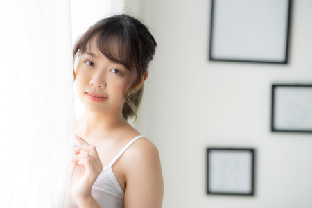 Portrait of beautiful young asian woman standing at the window and smile while wake up with sunlight at morning, girl happy with fresh and cheerful, lifestyle and relax for wellness concept.の写真素材