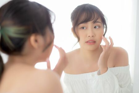 Beauty portrait young asian woman smiling look at mirror of checking skin care caucasian with wellness in the bedroom, beautiful girl happy touching face in reflection for health, lifestyle concept.の写真素材