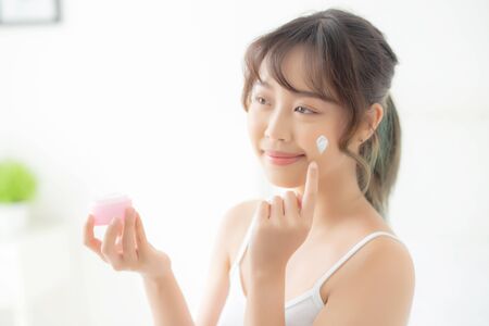 beautiful young asian woman happy applying cream or lotion with moisturizer to skin face, beauty asia girl applying skincare touch facial with cosmetic makeup, healthy and wellness concept.の写真素材