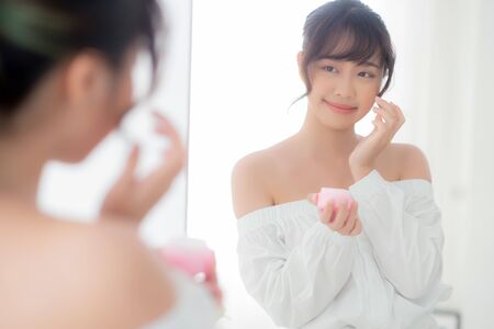 Beautiful portrait young asian woman applying cream moisturizer or lotion skin care cosmetic on face looking mirror, girl with treatment facial, health and wellness concept.の写真素材