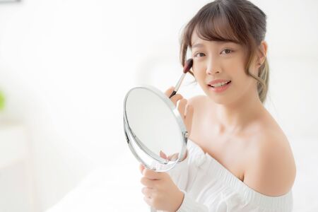 Beauty portrait young asian woman smile with face looking mirror applying makeup with brush cheek in the bedroom, beautiful of girl holding blusher, skin care and cosmetic concept.の写真素材