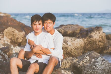 Homosexual portrait young asian couple sitting hug together on rock or stone in the beach in summer,...